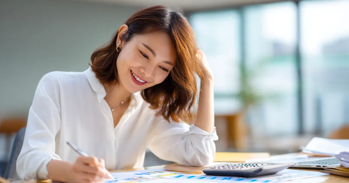 Hình ảnh mô phỏng người phụ nữ ghi chép bên bàn làm việc có máy tính cầm tay và bảng biểu
